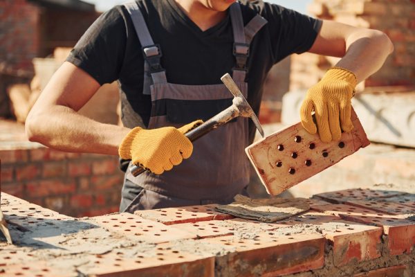 Holding brick and using hammer. Construction worker in uniform and safety equipment have job on building.